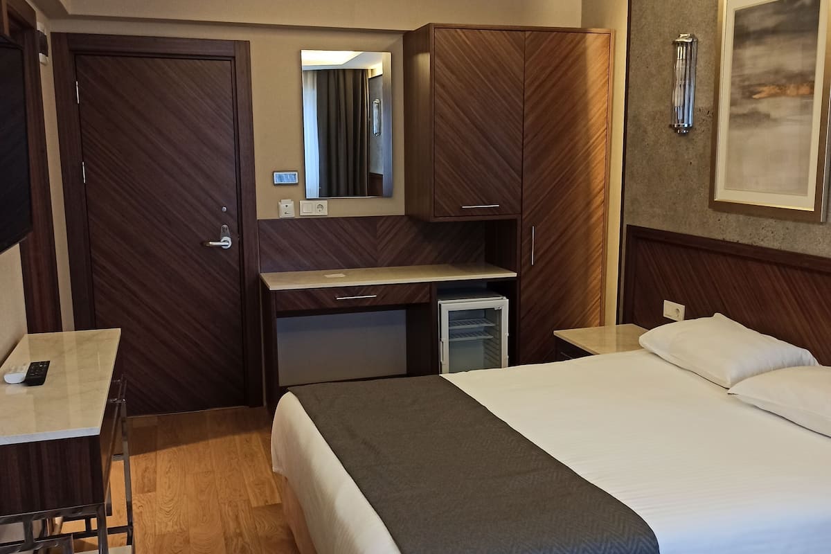 A hotel room with a bed, a desk, a chair, a wardrobe, a mirror, and a painting on the wall.