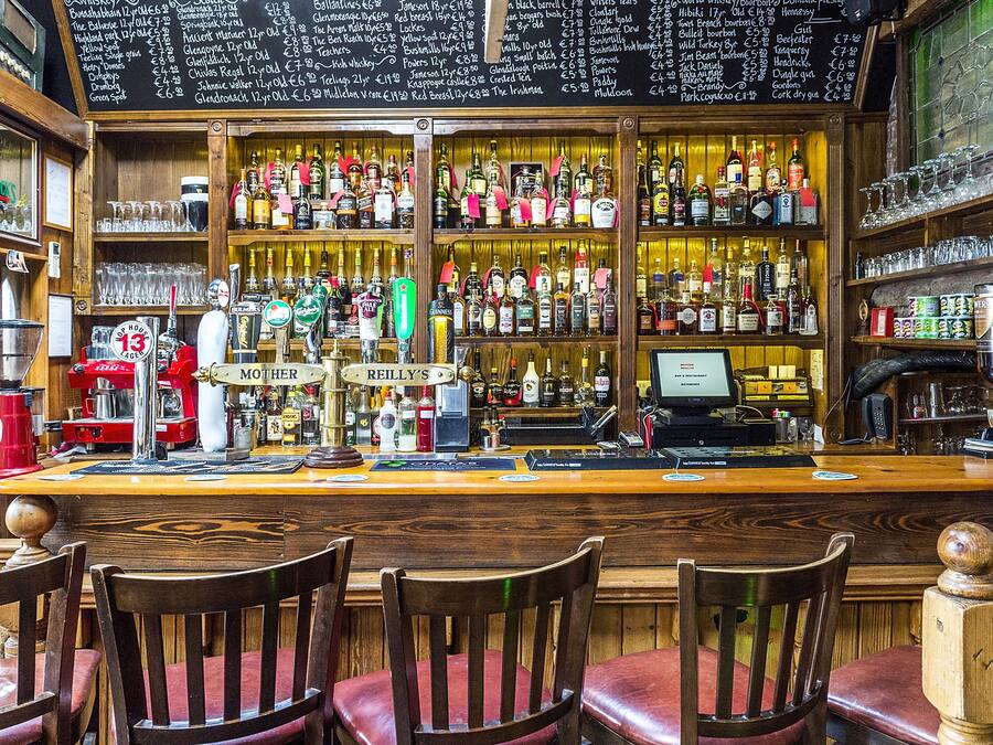 A bar with wooden shelves stocked with various bottles and glasses, a cash register, and a chalkboard menu.