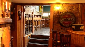 Un área de bar con mobiliario de madera, una pared de piedra con un letrero y una escalera que lleva a otra habitación.