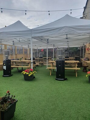 A covered outdoor area with multiple wooden picnic tables and artificial green turf. - The Uppercross House Hotel (Dublin)