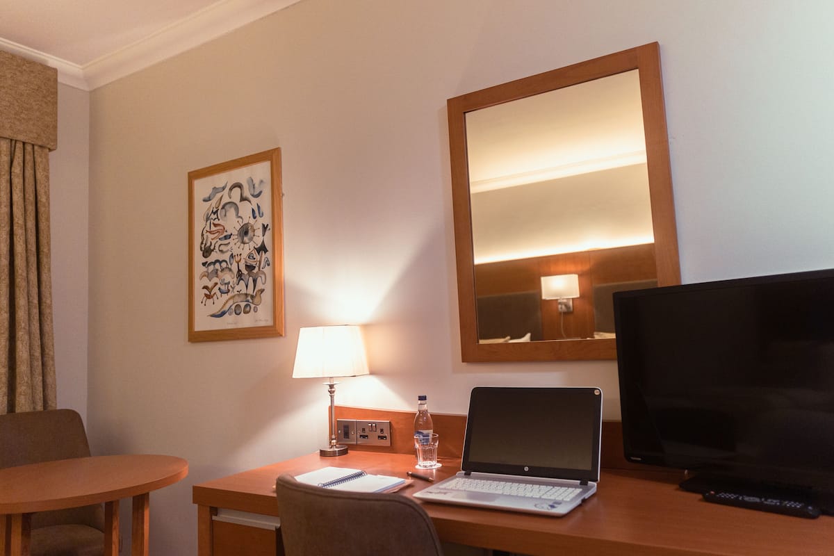 Ein Hotelzimmer mit einem Holztisch, einem Laptop, einem Fernseher, einer Lampe, einer Wasserflasche und einem gerahmten Kunstwerk an der Wand.