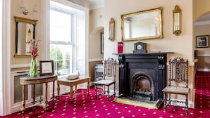 Una habitación con chimenea, sillas ornamentadas, un jarrón con flores rojas y una alfombra roja.