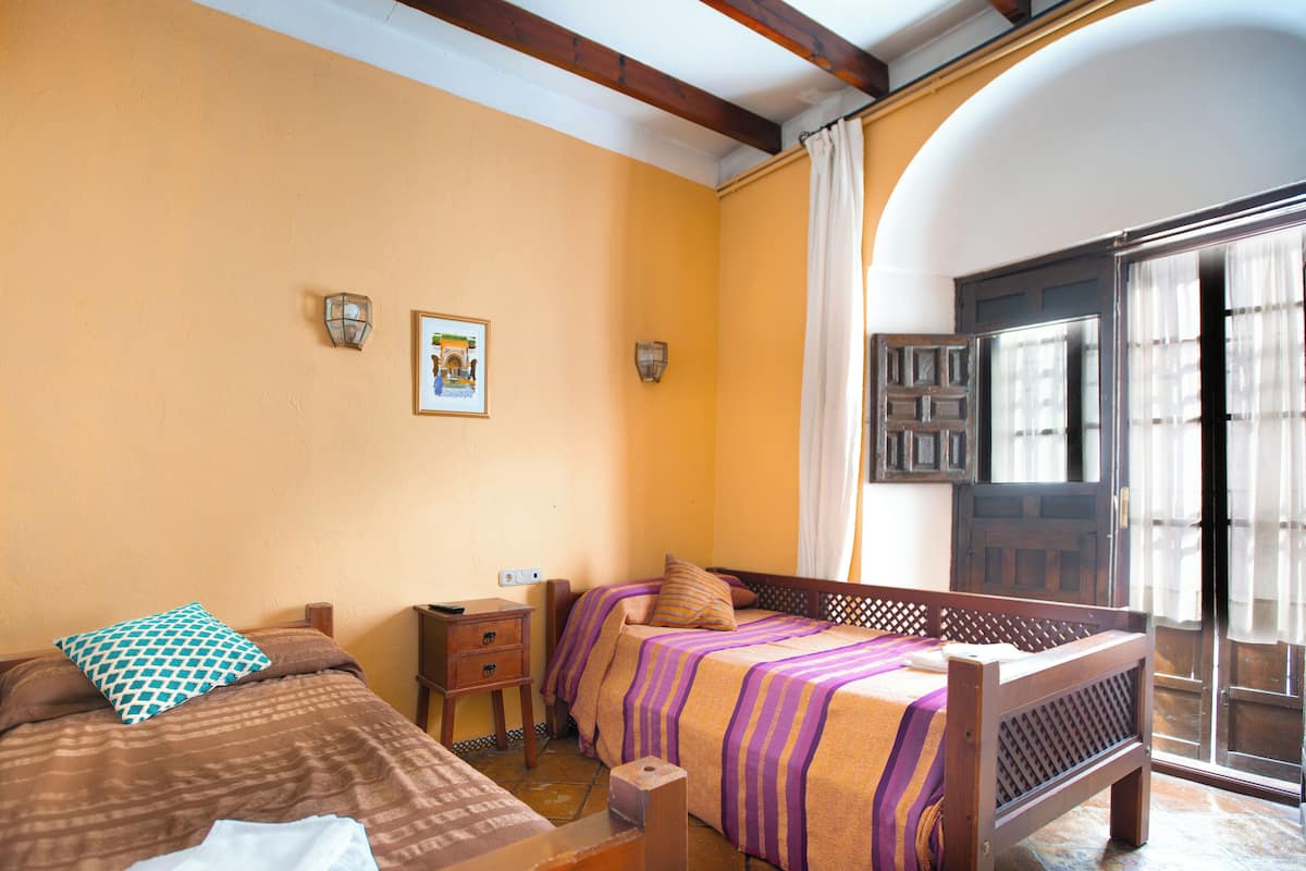 Una habitación con dos camas, una mesita de noche de madera y un ventanal con cortinas.