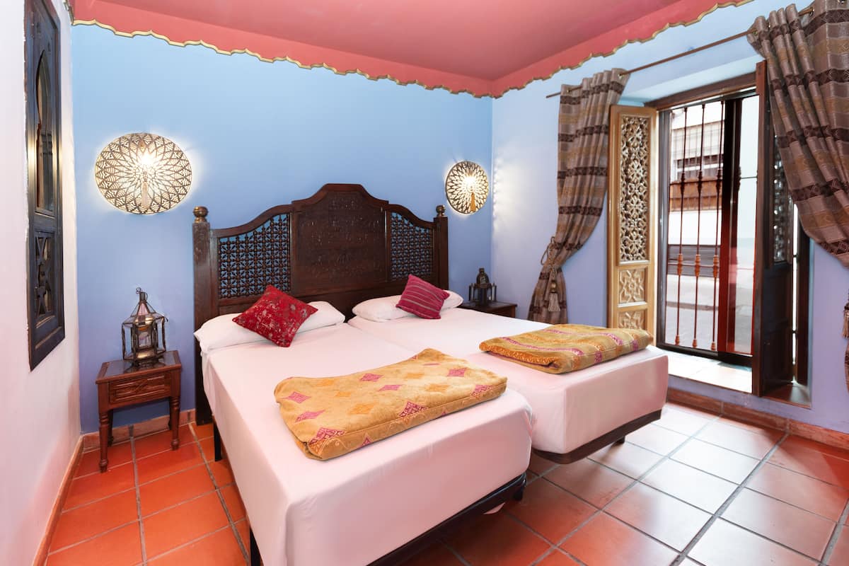 Un dormitorio con dos camas, una mesita de noche, apliques decorativos en la pared y vistas a través de una ventana con cortinas.
