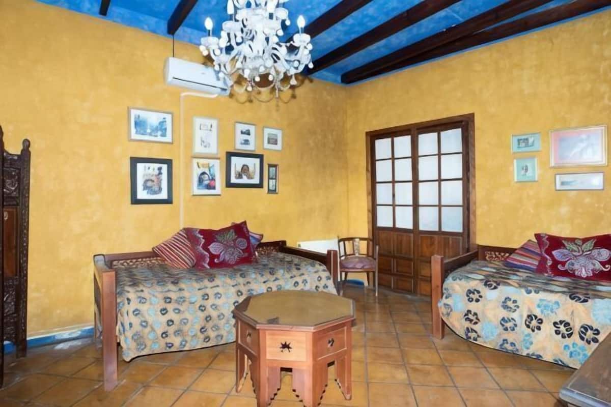 Una habitación con dos camas, una mesa de madera, un candelabro y un ventanal grande.