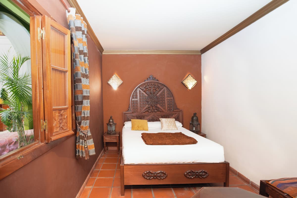 Un dormitorio con cama de madera, mesita de noche, lámpara de pared y ventana con cortina.