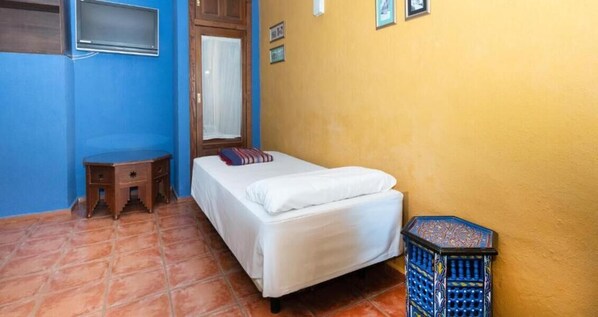 A small, single-bed room with a wooden nightstand, a window with a blind, and a tiled floor.