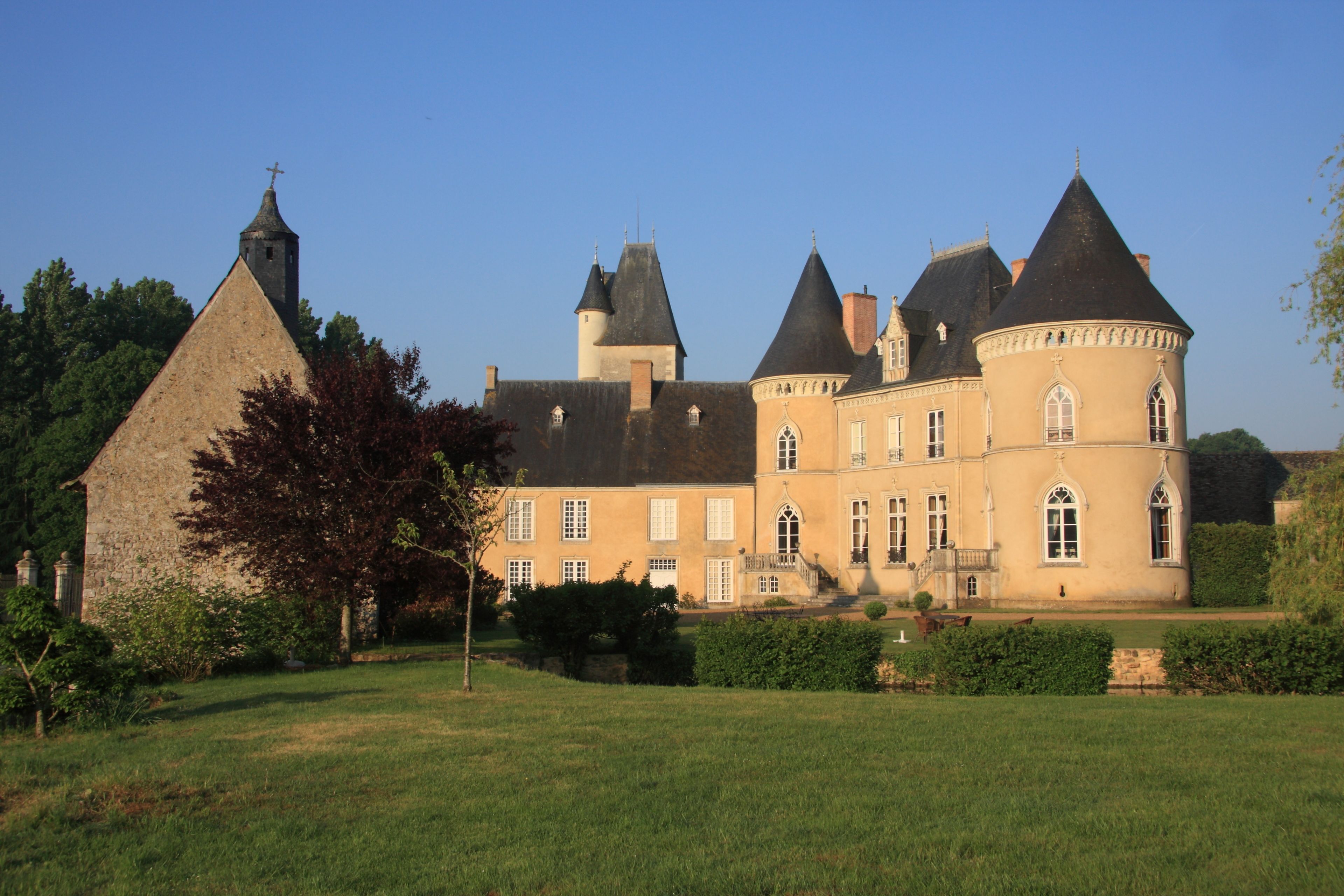 Un edificio grande e histórico con múltiples torres y un campanario destacado, rodeado de jardines bien cuidados y un cielo despejado.
