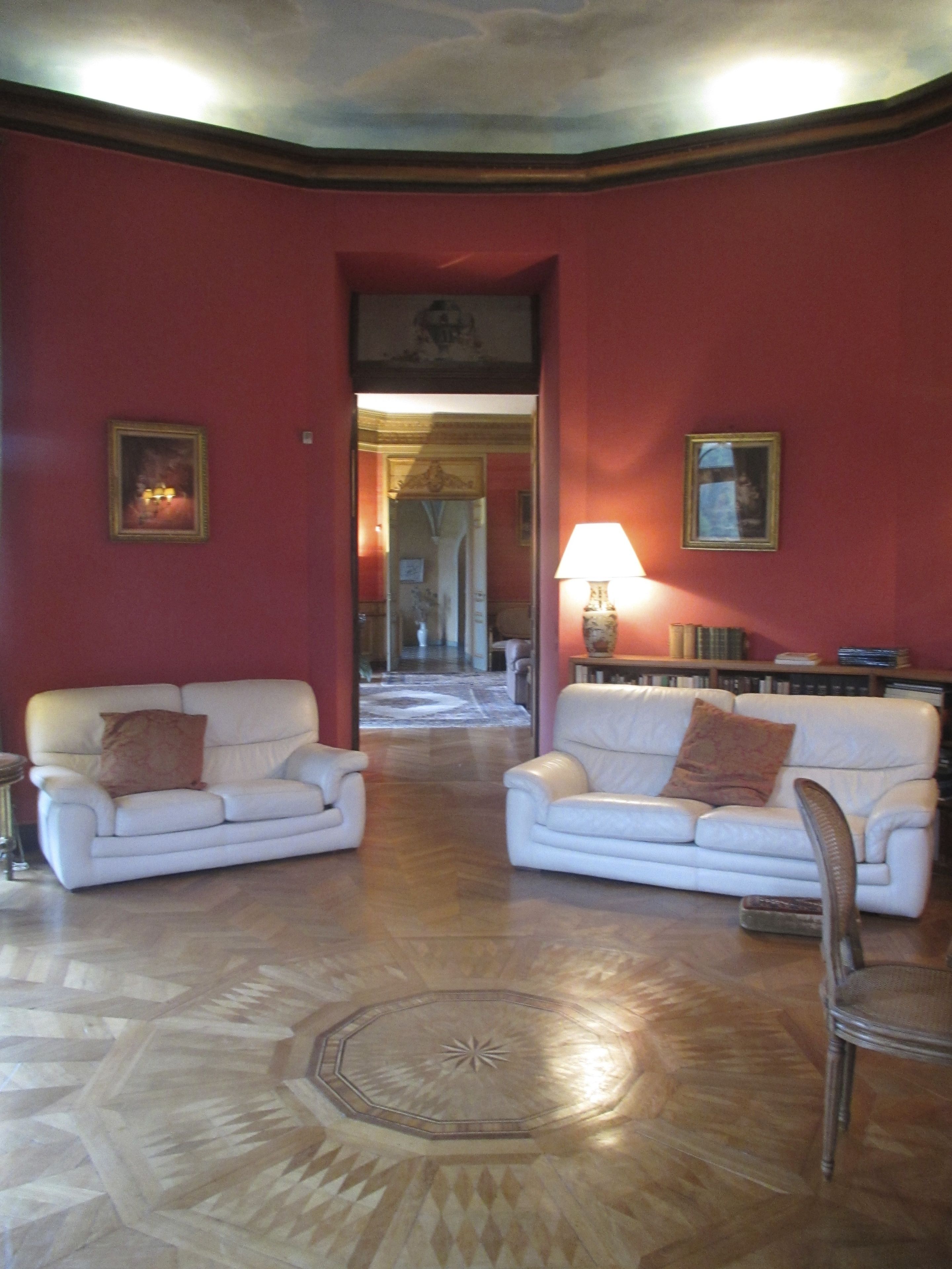 A room with two white sofas, a wooden floor with a geometric pattern, and a red wall with framed pictures.