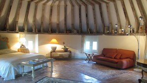 Una habitación con cama, sofá, mesa de centro, lámpara, lámpara de pie, una mesita con planta y una ventana con cortinas.