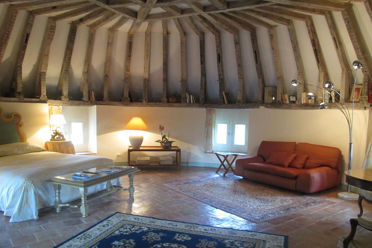 Una habitación con cama, sofá, mesa de centro, lámpara, lámpara de pie, una mesita con planta y una ventana con cortinas.