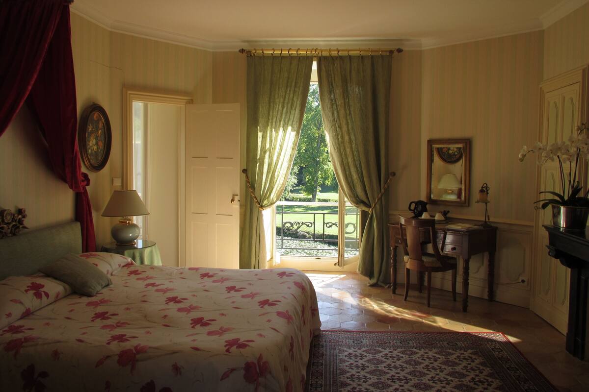 Un dormitorio con cama, escritorio, silla, lámpara, espejo y vistas al exterior.