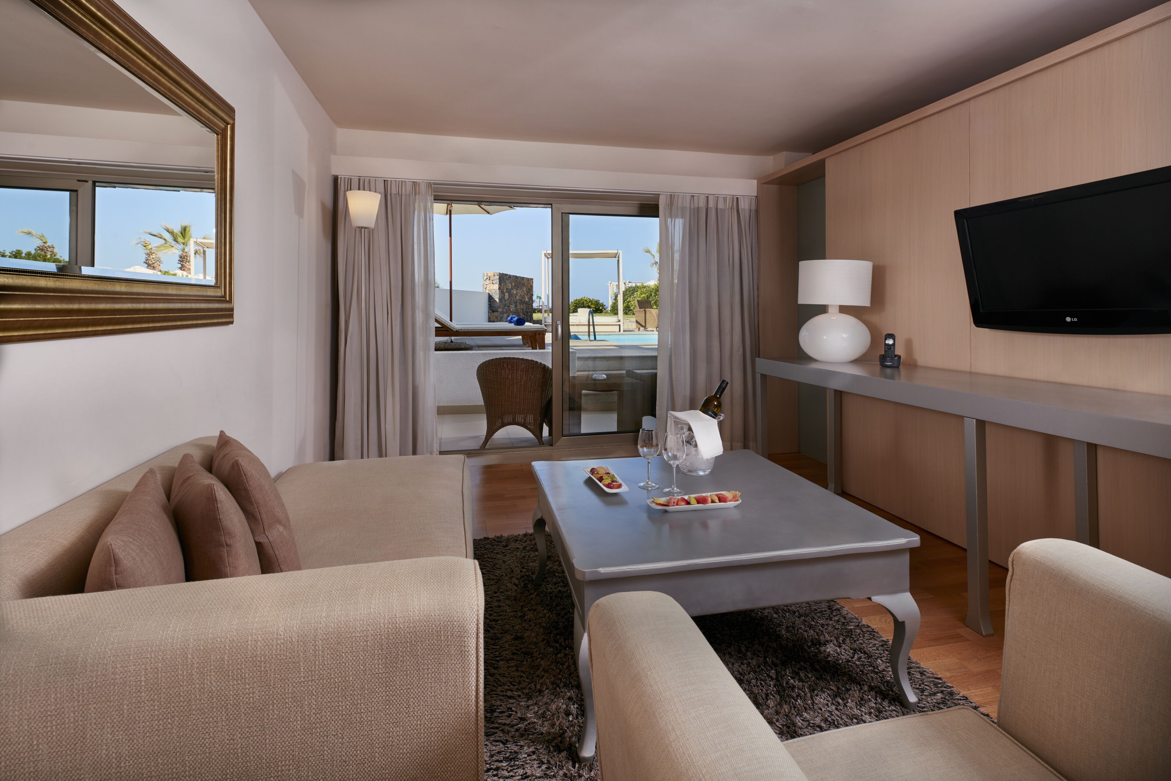 A hotel room with a sofa, a coffee table, a TV, and a view of the pool and palm trees.