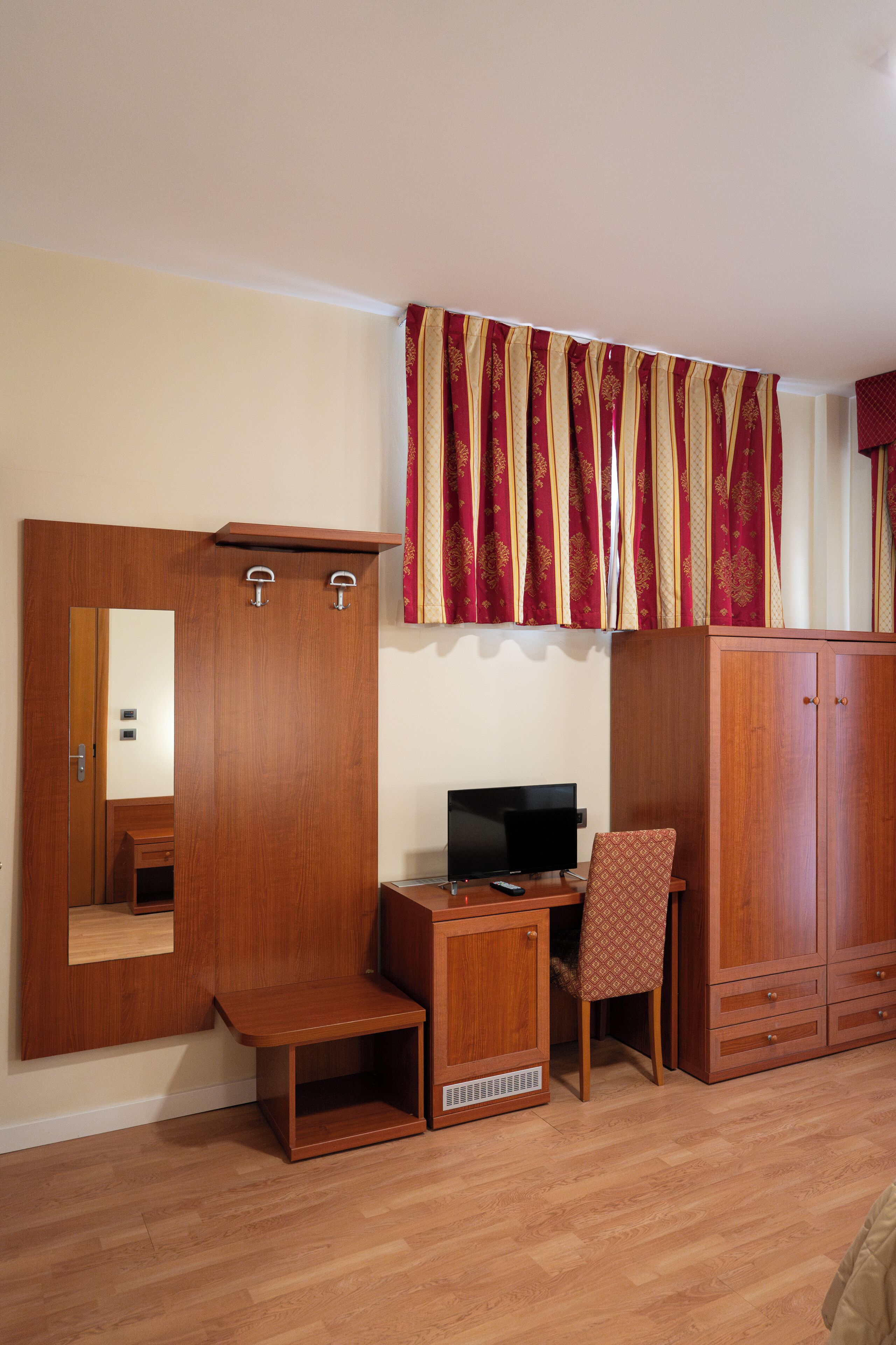 A room with a wooden wardrobe, a desk with a computer, a chair, and a mirror.