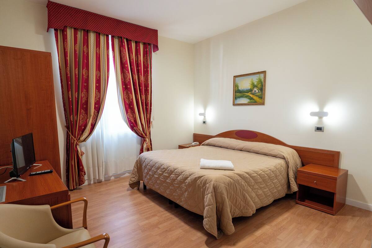 Ein Hotelzimmer mit Bett, Nachttisch, Lampe, Schreibtisch mit Computer, Stuhl und Vorhängen.