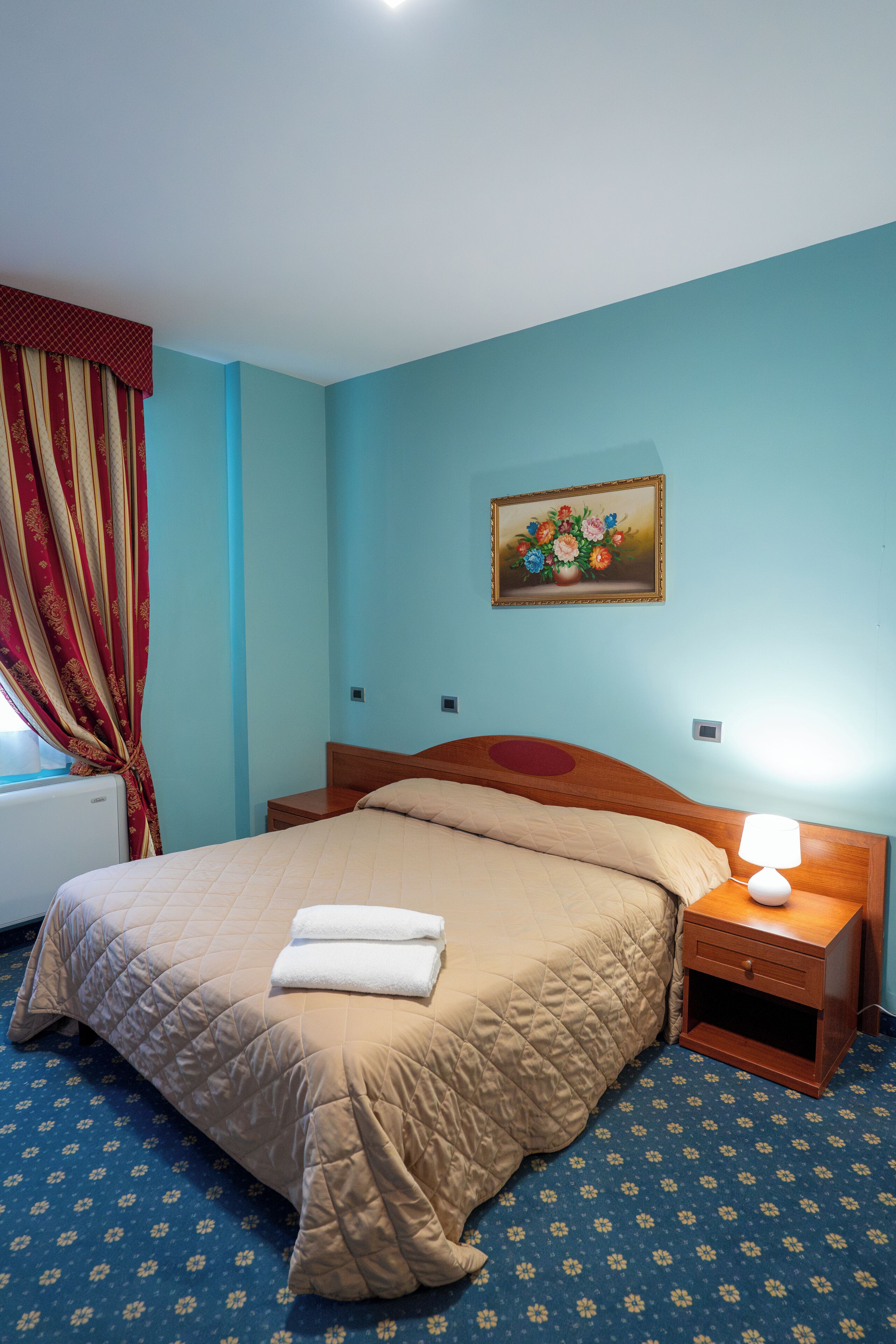 Een slaapkamer met een bed, een nachtkastje, een lamp, een schilderij aan de muur en een raam met gordijnen.
