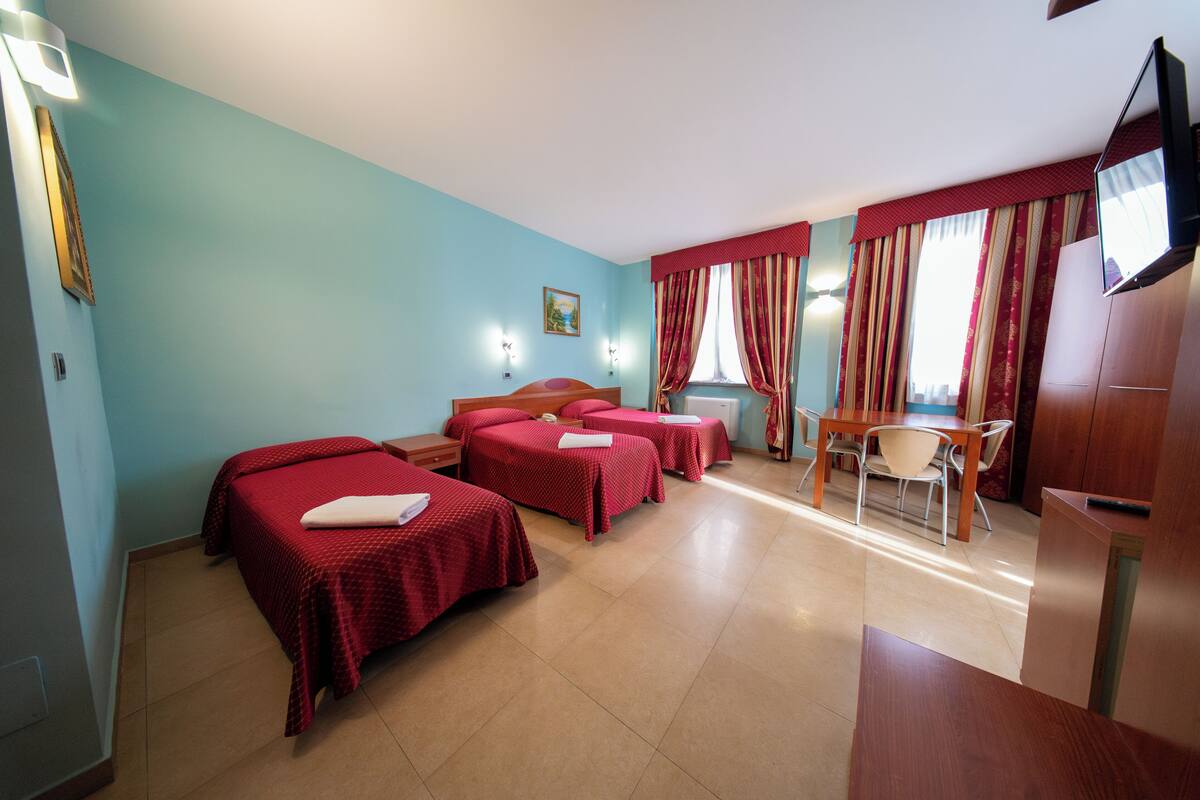 Ein Hotelzimmer mit vier Einzelbetten, einem Holztisch, einem Stuhl, einem Fernseher und einem Bild an der Wand.
