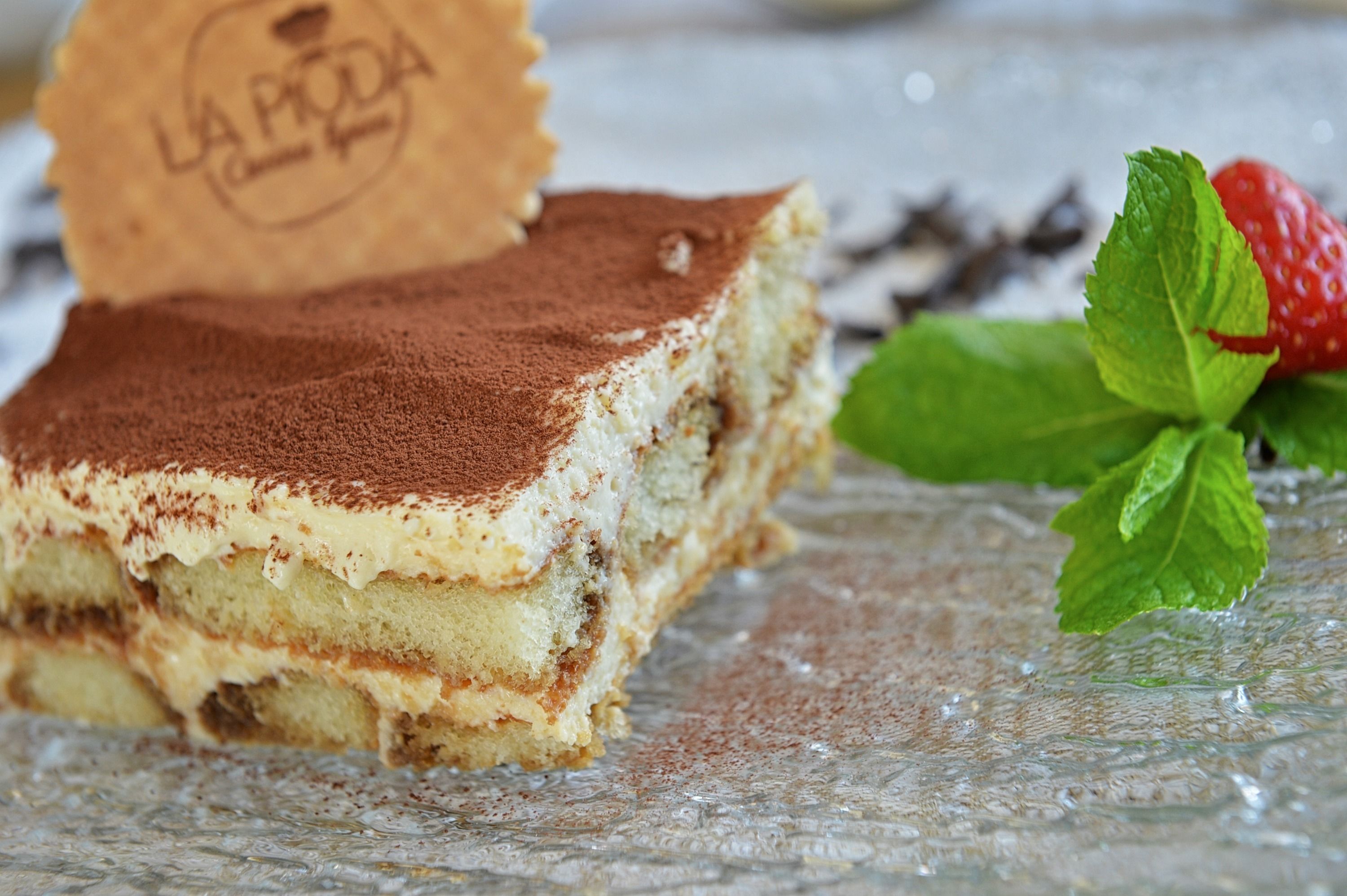 A slice of tiramisu on a glass plate with a mint leaf and a strawberry.