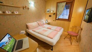 A bedroom with a bed, a desk with a laptop, a chair, a nightstand, and a window with curtains.