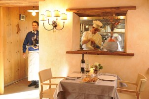 Interno di un ristorante con pareti in legno, un tavolo apparecchiato con piatti, bicchieri da vino e una bottiglia. Si intravede uno chef sullo sfondo.