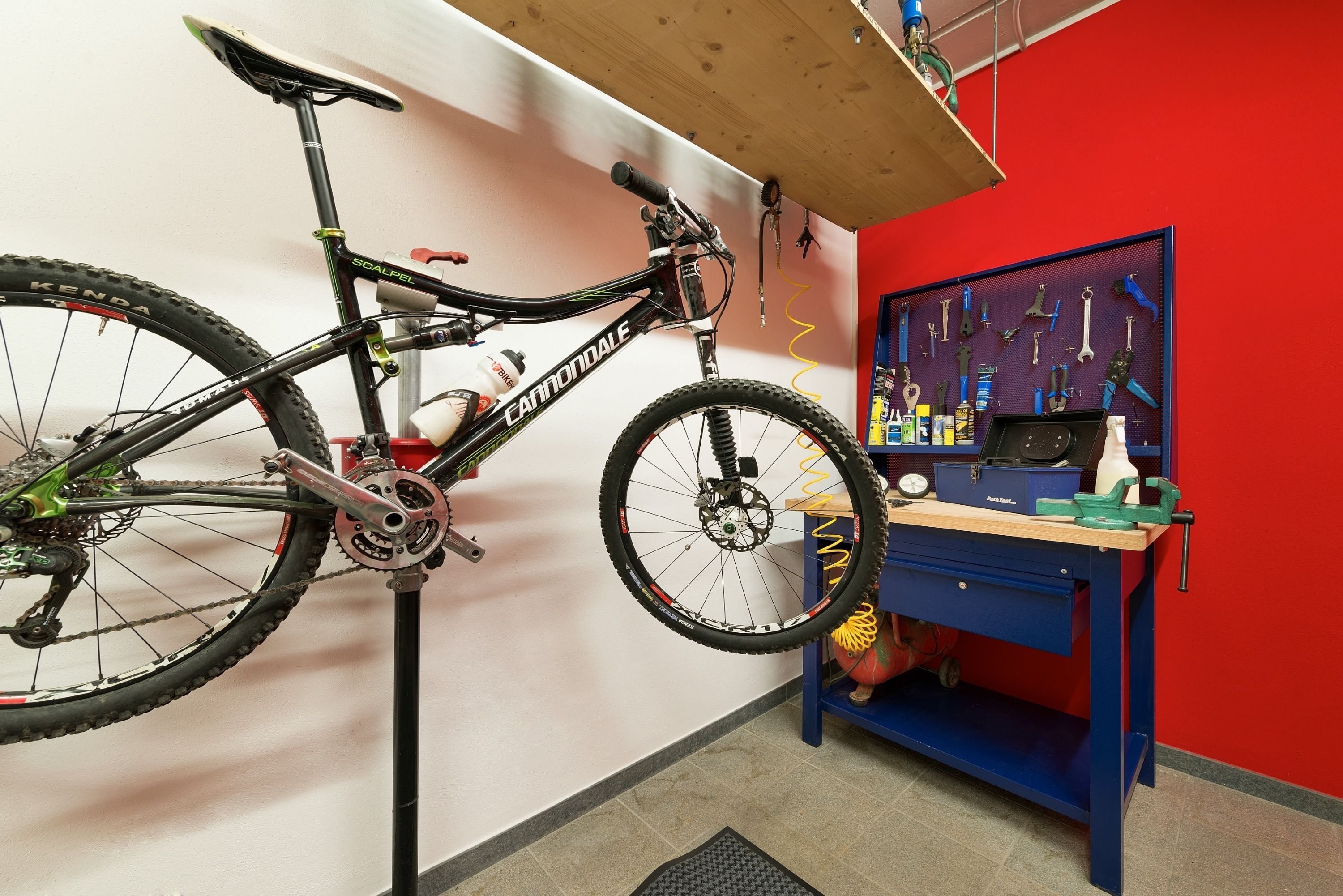 Un vélo est monté sur un support dans une pièce dont un mur est rouge, où l’on trouve également un établi bleu avec des outils, et un panneau perforé où sont accrochés d’autres outils.