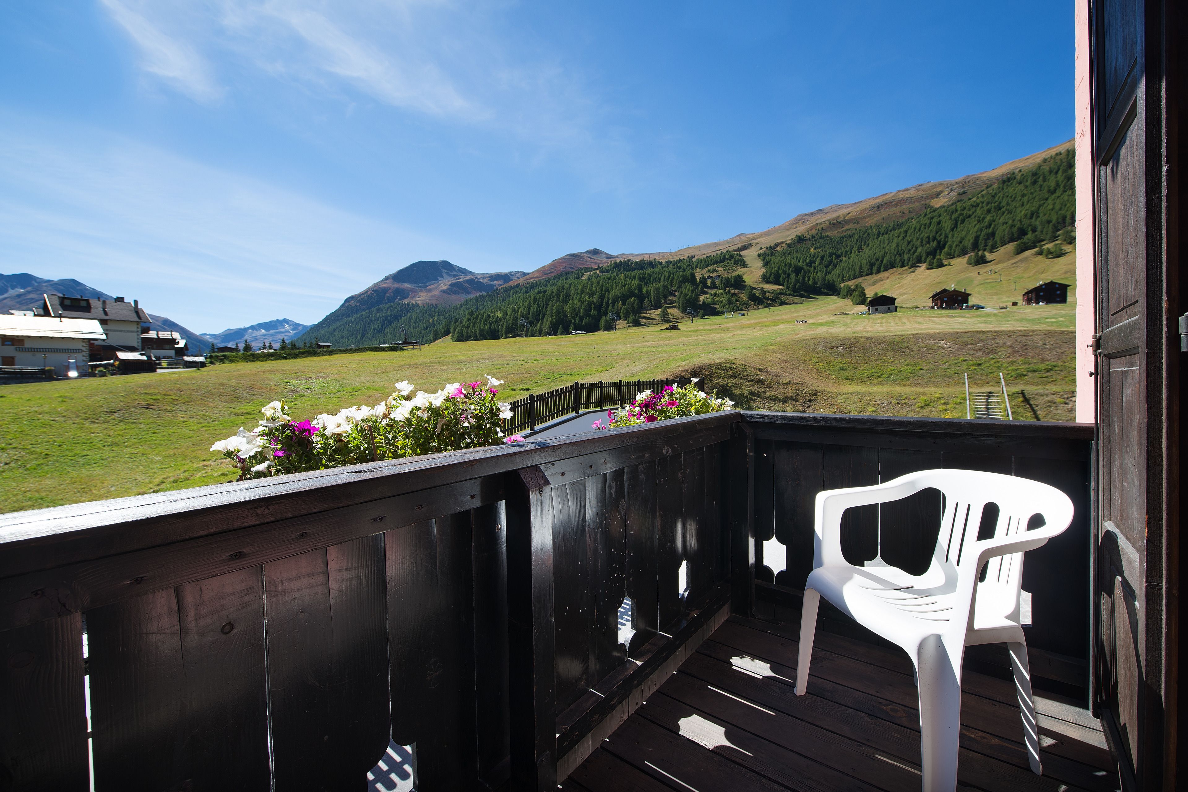 Una terraza de madera con una silla de plástico blanca, con vistas a un campo cubierto de césped y montañas al fondo.