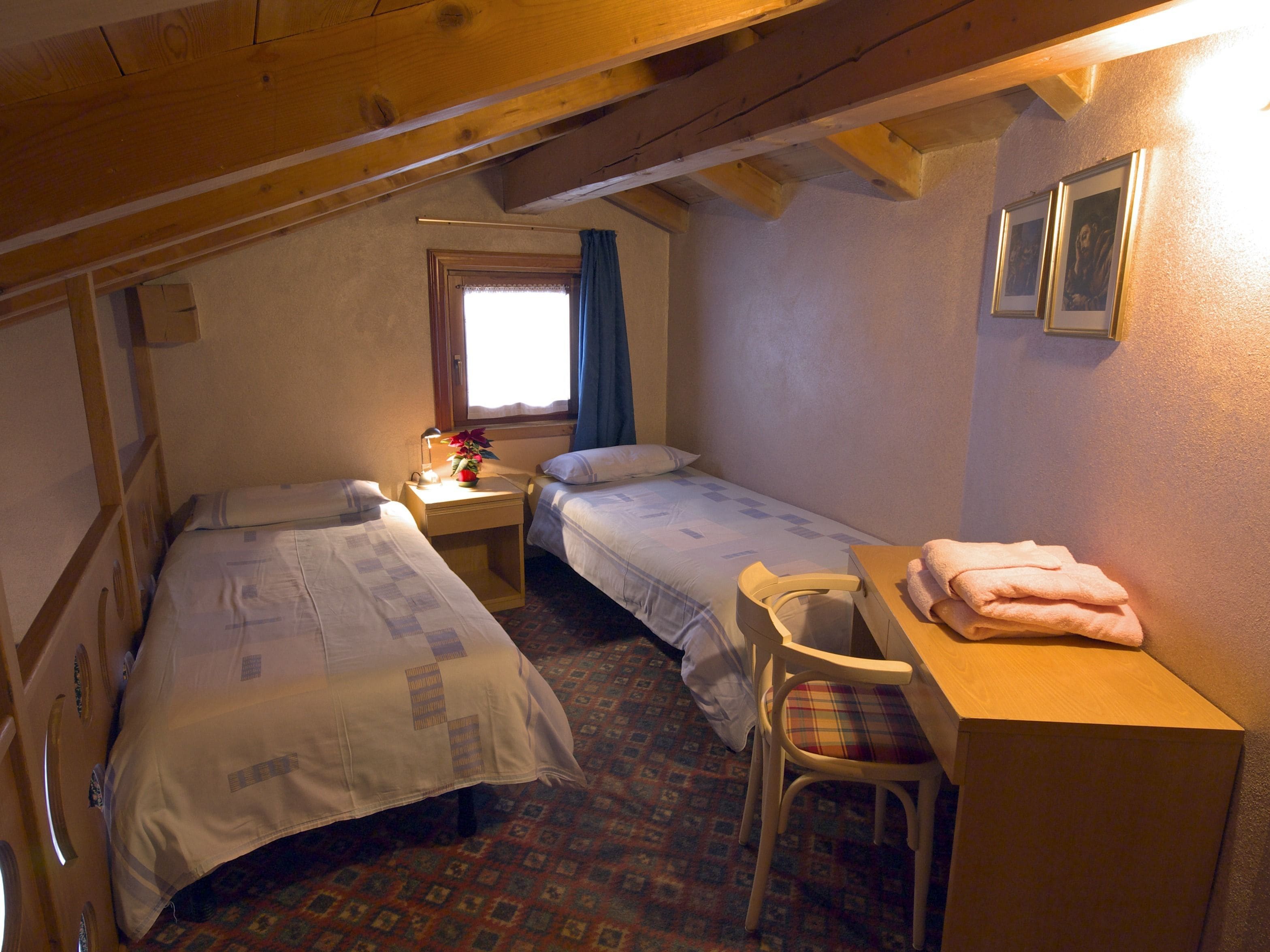 Una habitación pequeña y acogedora con dos camas individuales, un escritorio de madera con una silla, un ventanal con cortinas y un cuadro colgado en la pared.