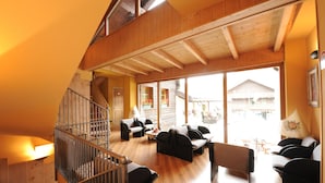 A spacious living area with a wooden staircase, multiple seating arrangements, and large windows offering a view of the outside. - Hotel Astra (Livigno)