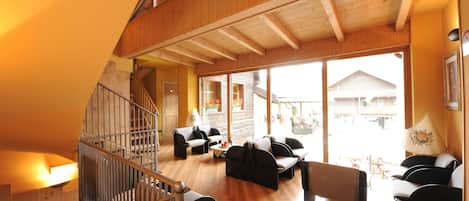 A spacious living area with a wooden staircase, multiple seating arrangements, and large windows offering a view of the outside.