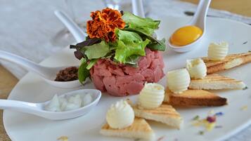 Une assiette contenant un plat à base de viande, agrémenté de feuilles vertes et d’une fleur, et accompagné de petits morceaux de beurre et de tranches de pain.