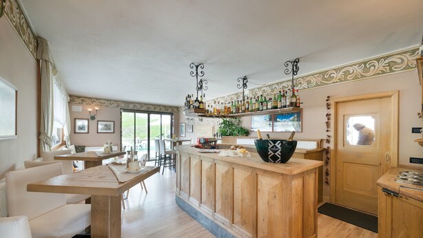 A bar area with wooden counters, a large green bowl, and a variety of bottles. There are white chairs and a dining area in the background. - Hotel Astra (Livigno)