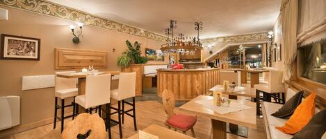 Un comedor acogedor y bien iluminado con mesas y sillas de madera, una zona de bar con mostrador y taburetes, y una planta grande en maceta.