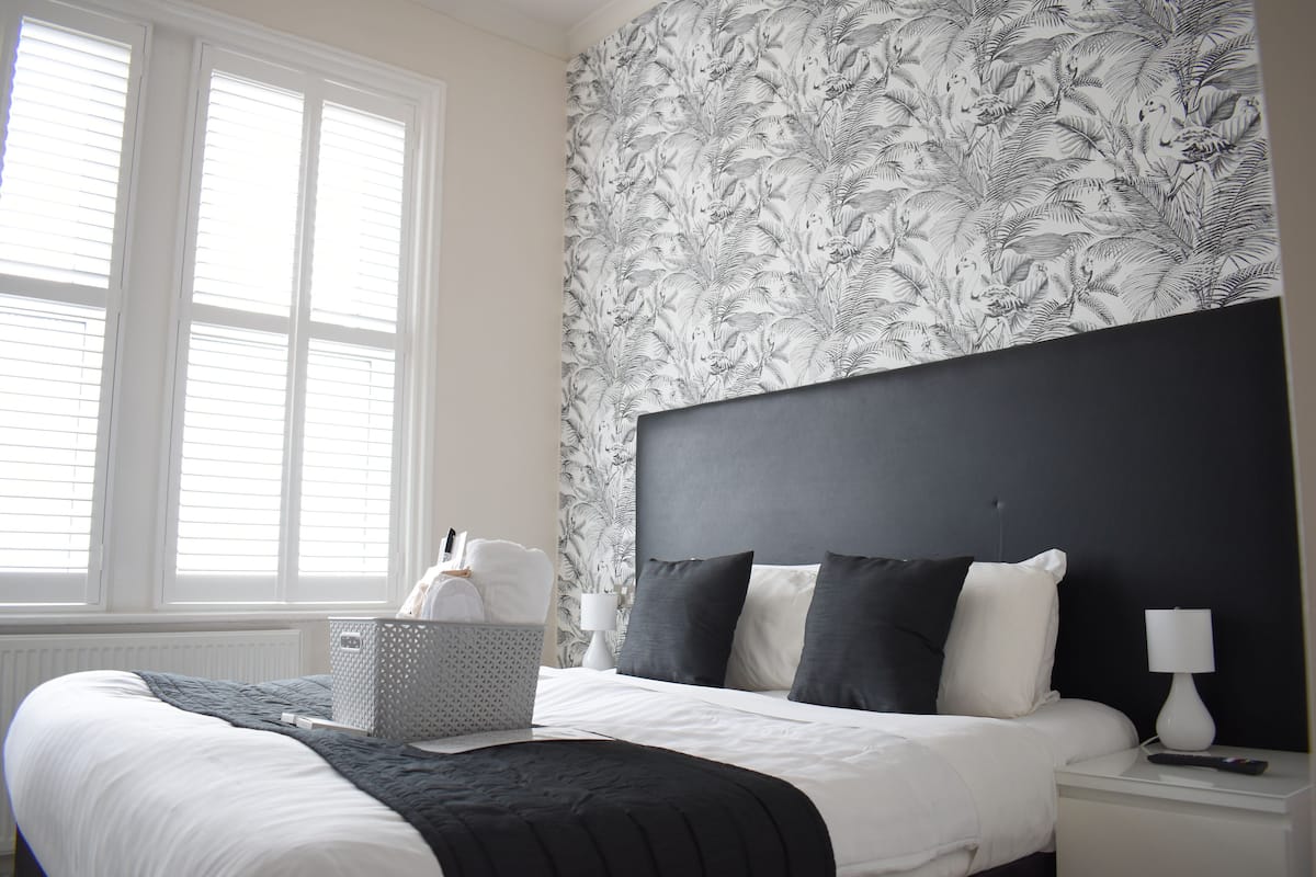 A neatly made bed with white and black bedding, a grey basket with towels, and a nightstand with a lamp and remote control.