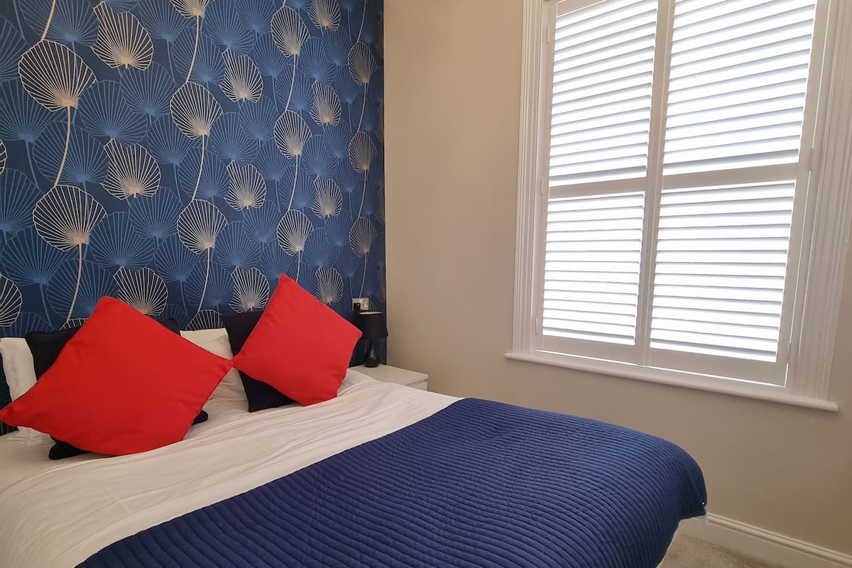 A bedroom with a bed, a blue and white patterned wallpaper, a red chandelier, and a window with blinds.