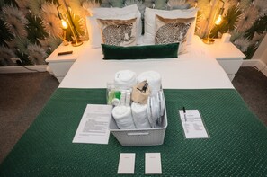Et pænt opredt hotelværelse med seng, puder, en kurv med toiletartikler og en grøn måtte.