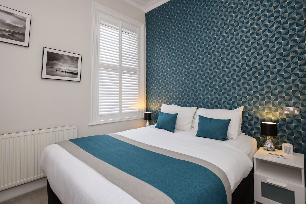 A neatly arranged bedroom with a bed, bedside tables, a radiator, and a window with blinds.