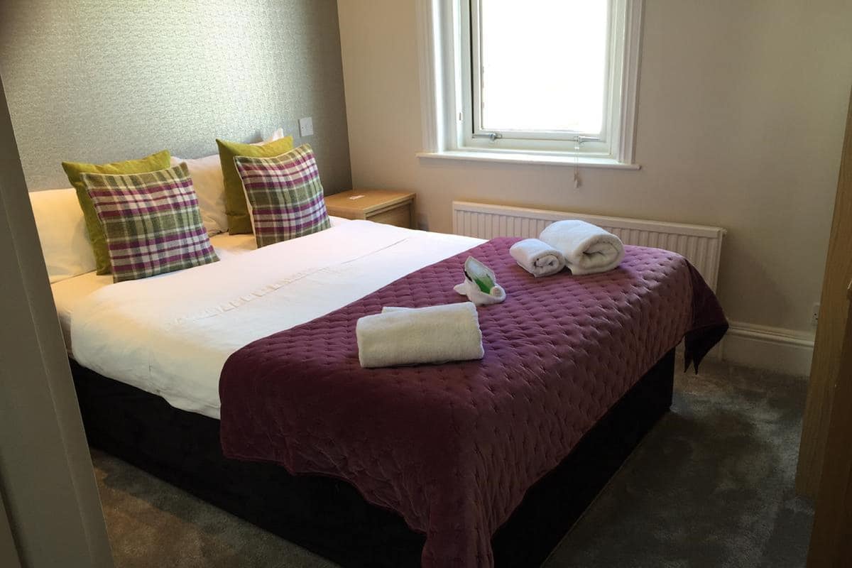 A neatly made bed with a purple bedspread, two pillows, and rolled towels.