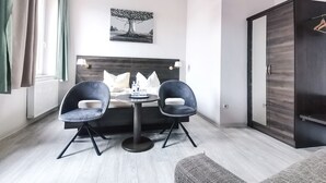 Una habitación de hotel moderna con cama, dos sillones, una mesa, una lámpara colgante, un cuadro y un armario.