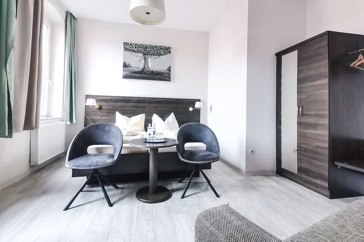A modern hotel room with a bed, two chairs, a table, a hanging light, a painting, and a wardrobe.