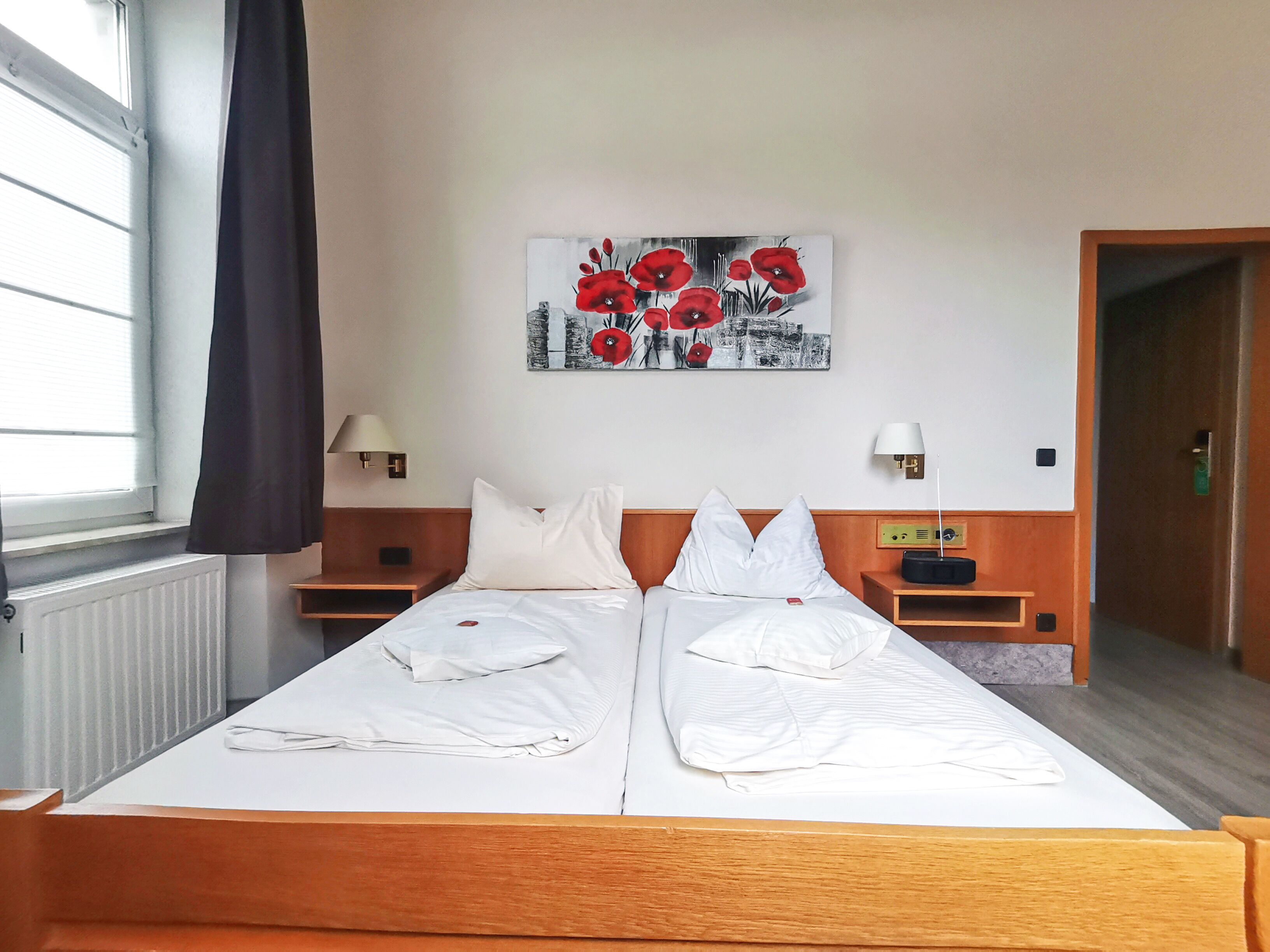 A double bed with white linens, two bedside tables with lamps, a wall-mounted artwork of red flowers, and a window with a black curtain.