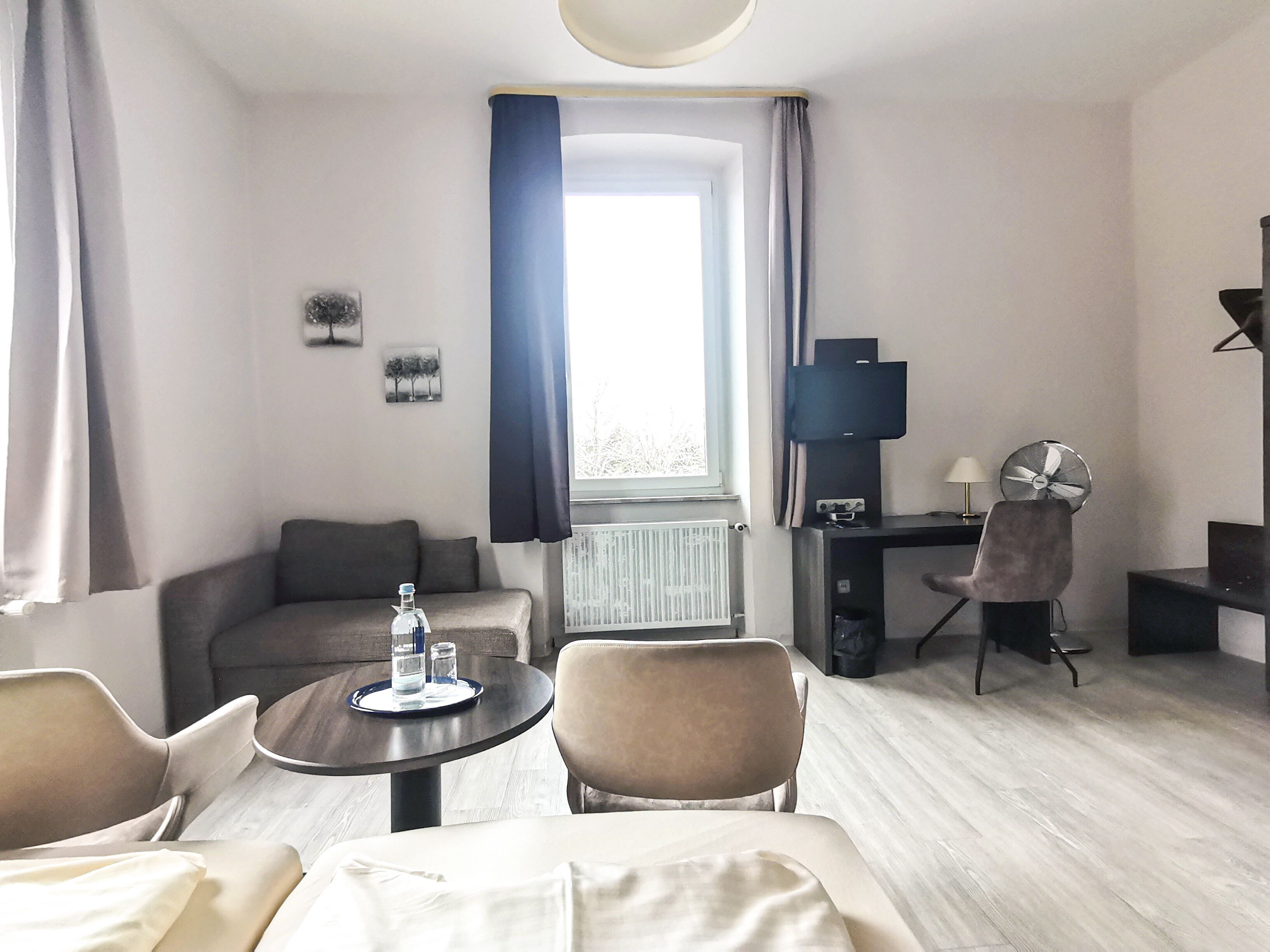 A modern hotel room with a bed, a desk, a chair, a small table with a bottle and glasses, a TV, and a window with curtains.