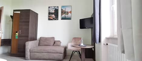 Habitación de hotel con sofá, una silla, una mesita con una botella de agua, televisor montado en la pared y dos cuadros enmarcados.