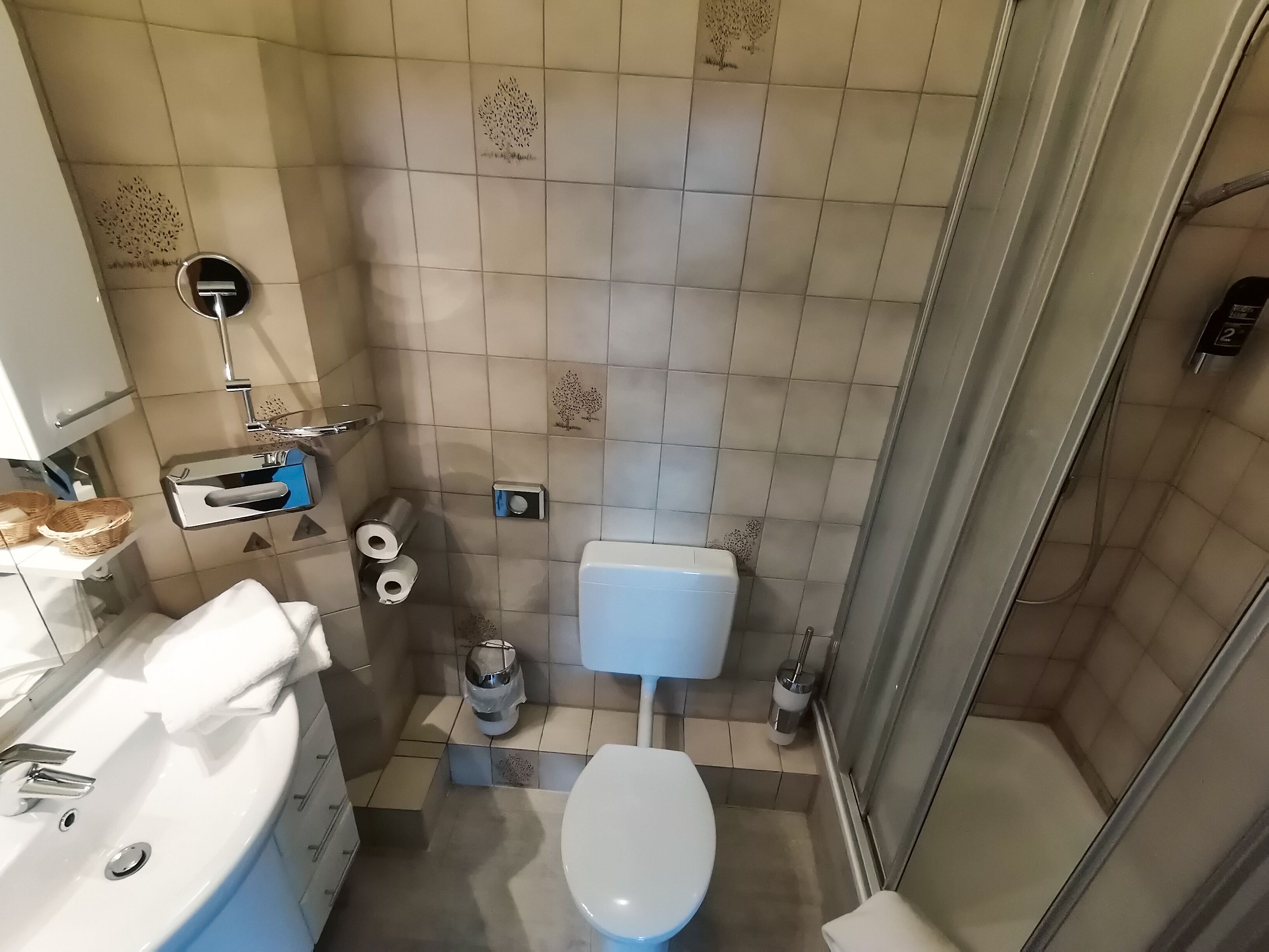 Une petite salle de bain avec des toilettes, un lavabo et une douche. On y trouve deux paniers sur le comptoir du lavabo, un porte-rouleau de papier toilette et une poubelle.