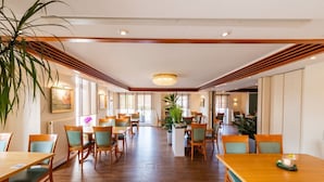 Un comedor amplio con mesas y sillas de madera, ventanas grandes y una planta en el centro.