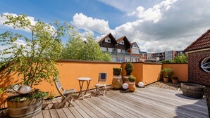 Una terraza de madera con mobiliario exterior, plantas en macetas y vistas a edificios residenciales.