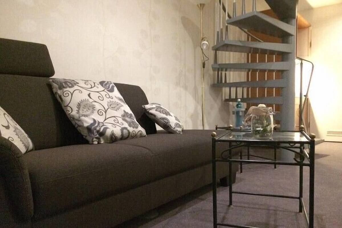 Un salon avec un canapé, des coussins à motifs, une table basse en verre et un escalier en arrière-plan.
