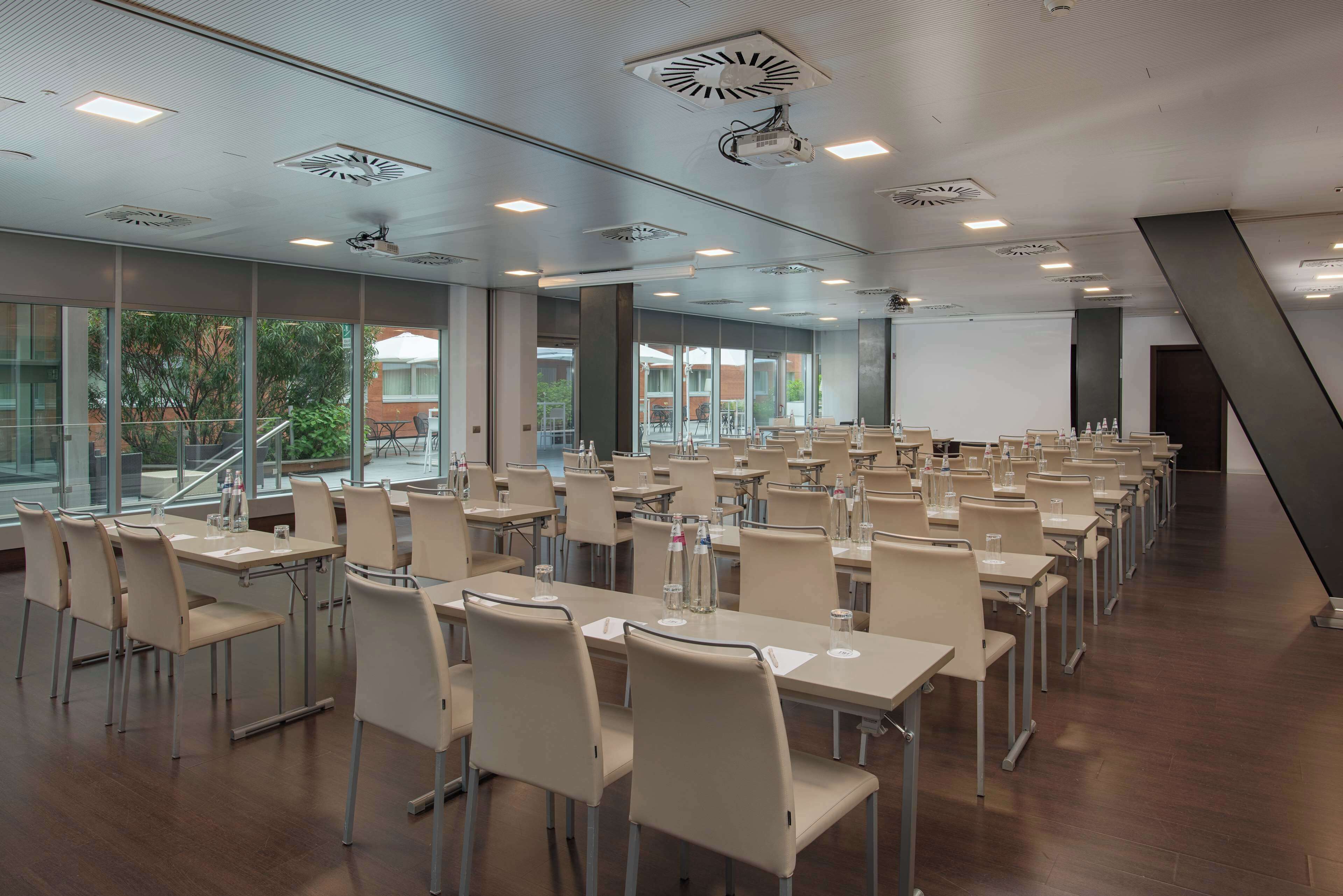 Ein Konferenzraum mit mehreren Tischen und Stühlen, großen Fenstern und einem Projektor an der Decke.