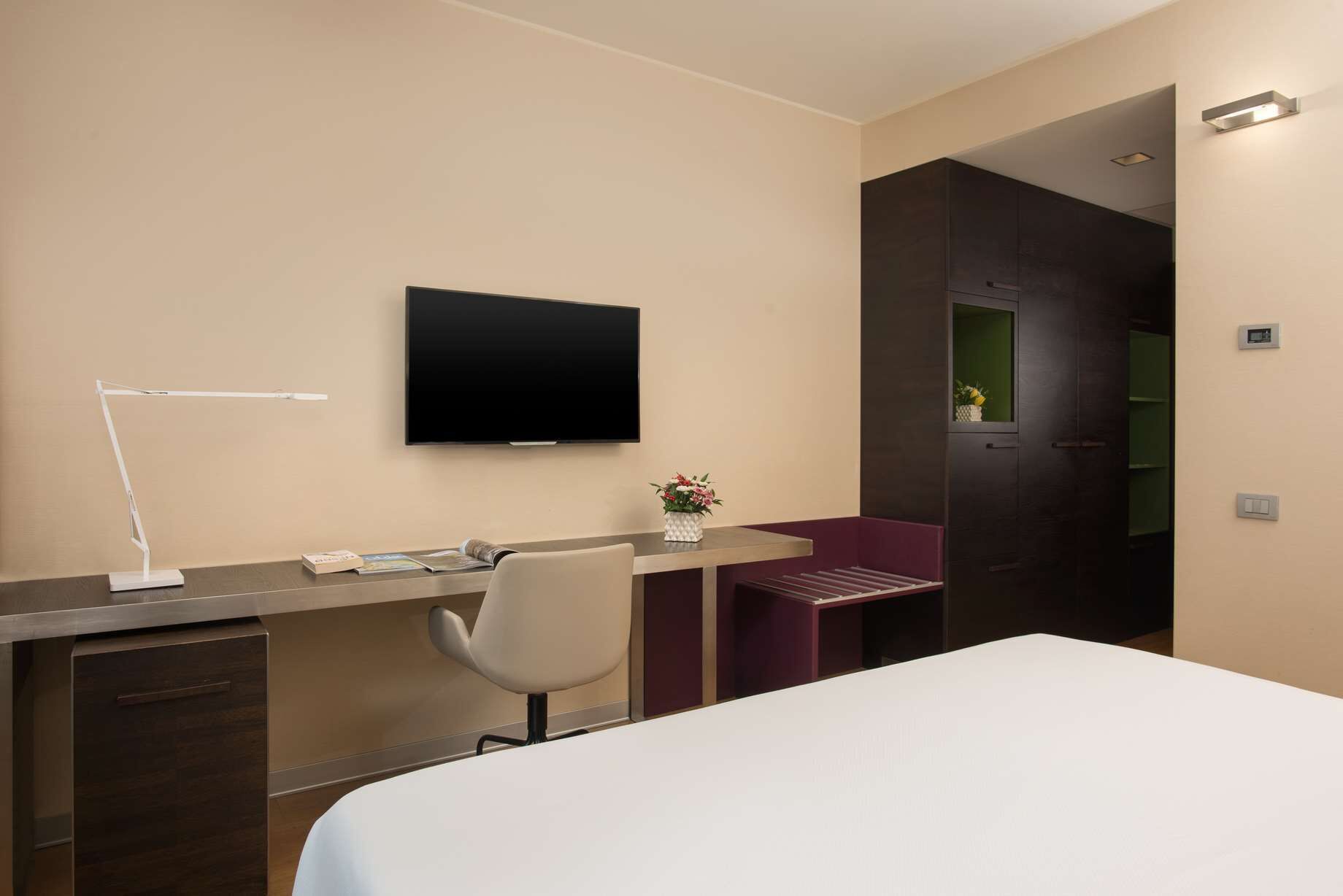 Una camera d'albergo con un letto, una scrivania con una sedia, una televisione e un tavolino con un vaso di fiori.