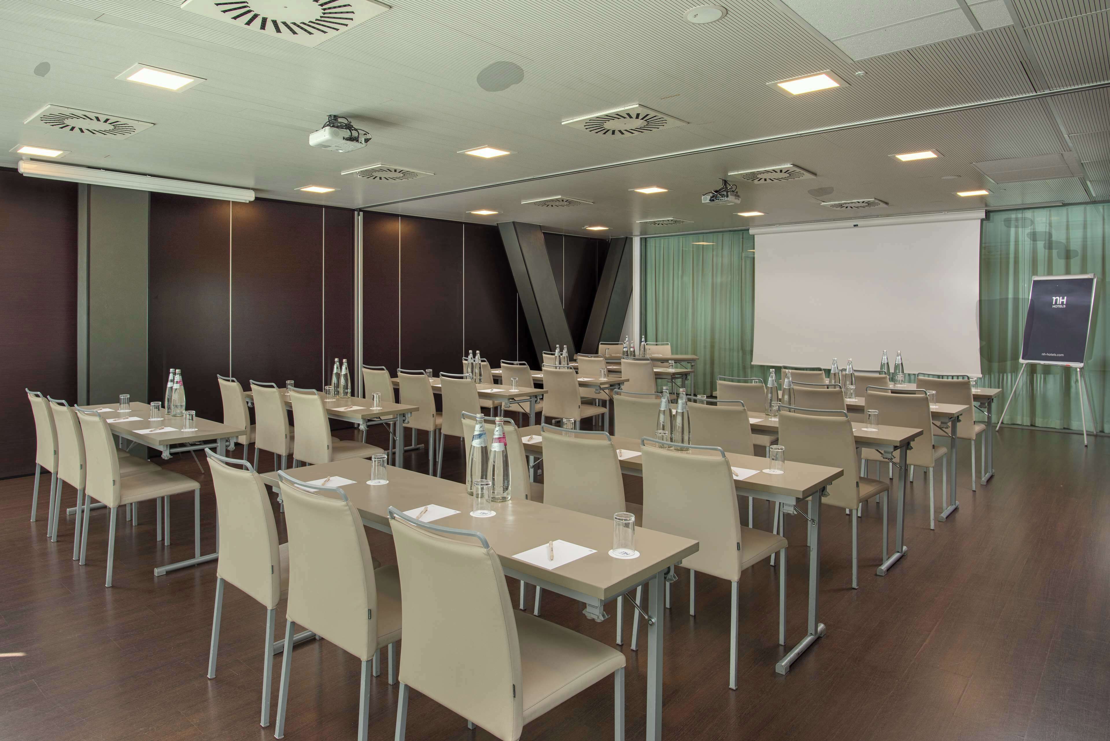 Un salón de conferencias con varias mesas y sillas, equipado con botellas de agua y vasos, y una pantalla para proyector.