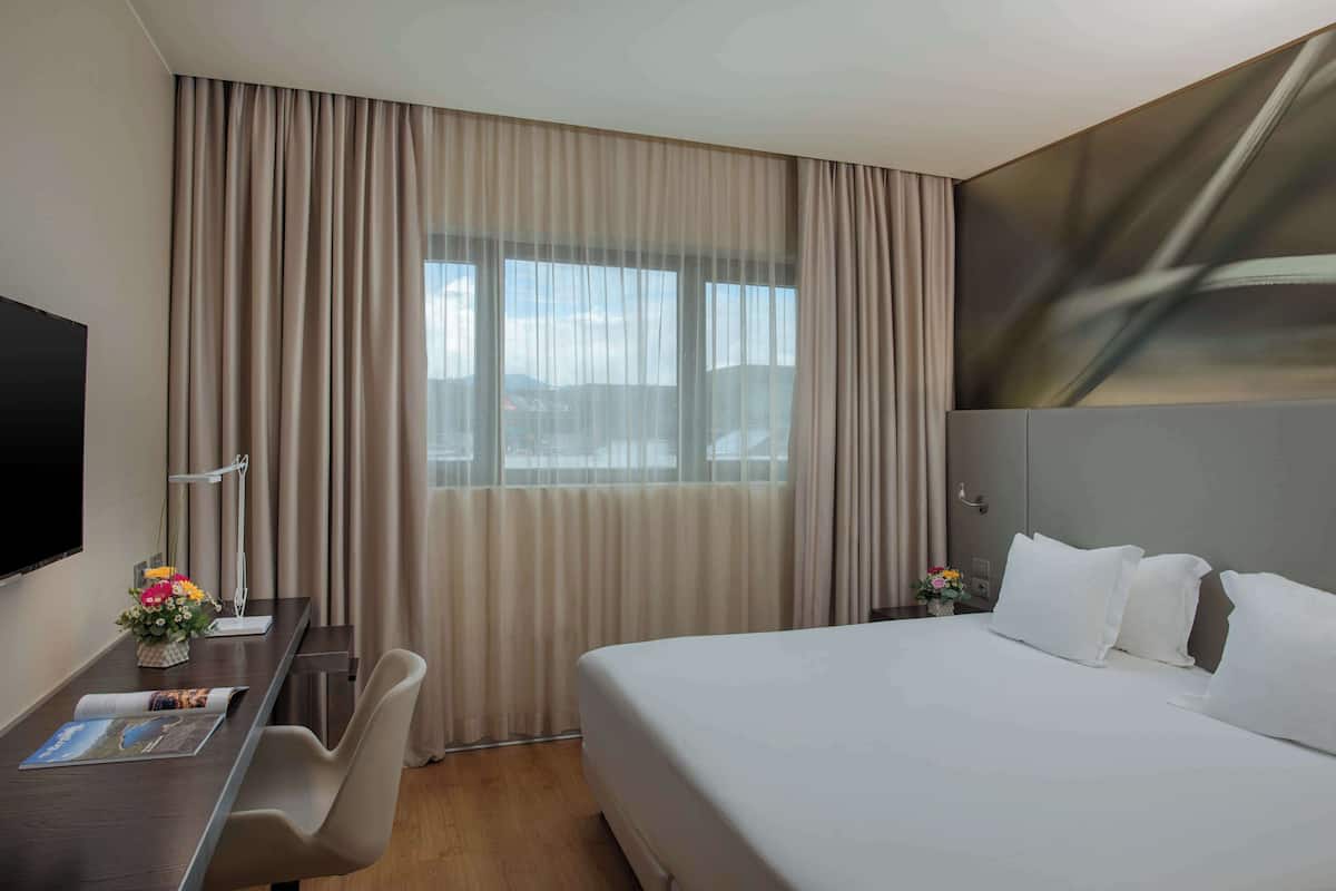 Ein Hotelzimmer mit einem großen Bett, einem Schreibtisch mit Stuhl, einem Flachbildfernseher und einem Fenster mit Vorhängen.